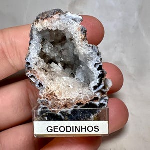 Op de afbeelding: Een geode-exemplaar met een kristalhelder interieur, gepresenteerd op een heldere plastic basis met het woord "GEODINHOS" erop gedrukt. De buitenkant van de geode is een mix van bruin, zwart en wit, terwijl de binnenkant fonkelt met heldere kristallen.