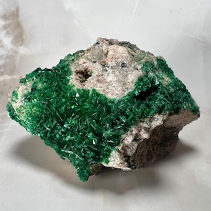 Op de afbeelding: Een natuurlijk mineraal specimen met een cluster van levendige, smaragdgroene kristallen. De kristallen zijn dicht opeengepakt op een ruwe, beige matrix. De algemene vorm is onregelmatig en toont de ruwe schoonheid van het mineraal. De achtergrond is een licht, gemarmerd oppervlak.