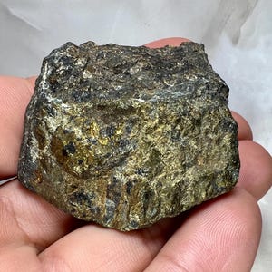 Pode incluir: Uma rocha áspera, de forma irregular, com brilho metálico. A superfície exibe uma mistura de cores cinza escuro, preto e dourado, com uma aparência texturizada e irregular. A rocha é segurada em uma mão, mostrando sua forma natural e bruta.
