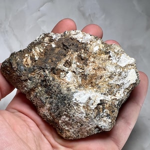 Pode incluir: Um espécime de rocha bruta e irregular apresentando uma mistura de depósitos minerais em tons de castanho terra, cinzento escuro e branco prateado brilhante. A superfície é texturizada e irregular, exibindo camadas cristalinas.