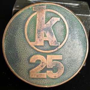 May include: A round, bronze-colored metal medallion with a green background. The medallion features a large letter "K" inside a circle, with the number "25" below. The metal has a textured surface.