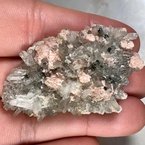 Peut inclure: Gros plan sur un spécimen minéral avec des amas cristallins transparents. Le spécimen présente des zones roses pâles et des inclusions noires, créant un aspect texturé. La palette de couleurs globale est neutre avec une touche de couleur.