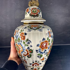 Può includere: Un vaso di ceramica bianco con coperchio, ornato da un motivo floreale arancione, blu e verde. Il coperchio è sormontato da una piccola scultura dorata. Il vaso è tenuto da una persona.