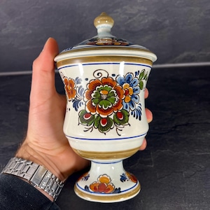 Könnte beinhalten: Ein weißes Keramikgefäß mit Deckel, verziert mit bunten Blumenmustern in Orange, Blau und Grün. Das Gefäß hat einen Sockel und einen goldenen Rand. Der Deckel ist mit einem kleinen, hellbraunen Knauf versehen.