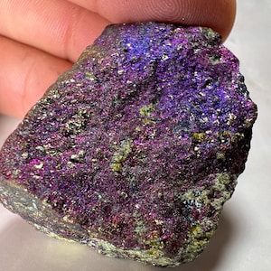 May include: A mineral specimen with a rough, iridescent surface. The rock displays a vibrant mix of colours, including deep purple, magenta, and blue, with touches of gold and green. The surface has a textured, crystalline appearance.