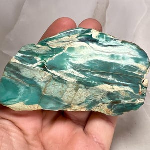 Può includere: Una pietra lucida, di forma irregolare, con un sorprendente motivo di tonalità verde acqua, bianche e marroni. La superficie della pietra mostra un effetto marmorizzato a strati, creando una trama visiva unica. La pietra naturale è tenuta in una mano.