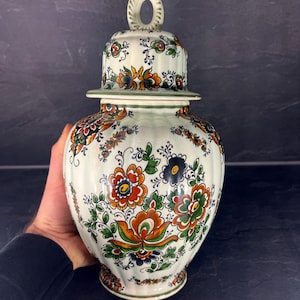 Può includere: Un vaso in ceramica color crema con coperchio, ornato con un motivo floreale arancione, verde e blu. Il vaso ha una forma bulbosa e una maniglia decorativa sul coperchio. Il design è ornato e dettagliato.