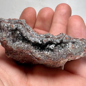 May include: A rough, natural mineral specimen with a cavity lined with sparkling crystals. The exterior is a mix of reddish-brown and gray, with the crystals appearing clear and reflective. The overall shape is irregular and organic.