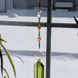 Puede incluir: Un atrapasoles decorativo colgado frente a una ventana. El atrapasoles presenta una cadena de cuentas coloridas en verde, naranja y amarillo, que conduce a un marco de metal cuadrado y un gran cristal rectangular verde. El fondo muestra una escena nevada al aire libre.