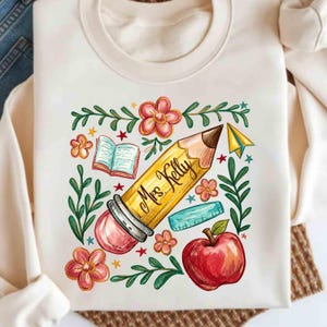 Peut inclure: Un sweat-shirt à col rond couleur crème arborant un motif central composé d'un crayon jaune avec le texte Mrs. Kelly écrit en écriture noire. Le design comprend une pomme rouge, un livre ouvert, une règle, des fleurs et des feuilles vertes.