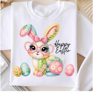 Peut inclure: Sweat-shirt blanc avec un lapin de dessin animé coloré portant des lunettes et un nœud. Le lapin est entouré d'œufs de Pâques décorés. Les mots "Happy Easter" sont écrits en script noir.
