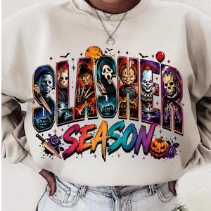 Peut inclure: Un sweat-shirt à col rond couleur crème avec une impression graphique portant le texte "SLASHER SEASON" en lettres colorées et stylisées. Le motif comprend des illustrations de divers personnages de films d'horreur, un couteau, une citrouille et des chauves-souris.