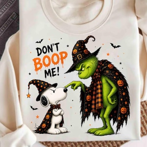 May include: A cream-colored sweatshirt featuring a graphic of a green creature and a white dog wearing matching black witch hats and capes with orange pumpkin patterns. The text DON'T BOOP ME! is printed in orange and black above the characters.