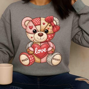 Peut inclure: Un sweat-shirt gris avec un motif d'ours en peluche en patchwork. L'ours tient un cœur rouge avec le mot "love" et porte un nœud rouge. Les pattes de l'ours disent "hugs" et "kisses".