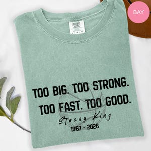 May include: A sage green short-sleeve t-shirt featuring black text that reads: TOO BIG. TOO STRONG. TOO FAST. TOO GOOD. Stacey King 1967-2026. A faint black outline of a basketball is printed behind the text. The shirt has a Comfort Colors brand label.