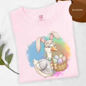 May include: A light pink t-shirt featuring a cartoon goose wearing a bunny mask and holding a basket of Easter eggs. The shirt has the words "Comfort Colors" on the tag. The background has a watercolor effect with blue, green, and purple hues.