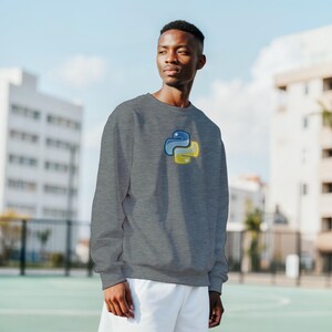 K&ouml;nnte beinhalten: Ein graues Sweatshirt mit dem Logo der Programmiersprache Python in Blau und Gelb. Das lang&auml;rmlige Rundhals-Shirt wird im Freien getragen. Das Python-Logo befindet sich auf der oberen linken Brustseite.