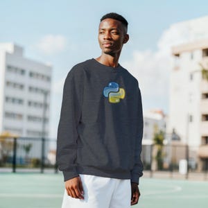 K&ouml;nnte beinhalten: Dunkelgraues Sweatshirt mit dem Python-Logo in Blau und Gelb. Das Sweatshirt ist ein lang&auml;rmeliger Rundhalsausschnitt. Die Person auf dem Bild tr&auml;gt wei&szlig;e Shorts.