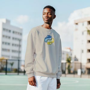 K&ouml;nnte beinhalten: Beiges Sweatshirt mit dem Python-Logo in Blau und Gelb. Das Sweatshirt ist ein Rundhals-Modell und wird von einer Person getragen. Der Hintergrund zeigt Geb&auml;ude und einen blauen Himmel.