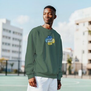 K&ouml;nnte beinhalten: Gr&uuml;nes Sweatshirt mit blau-gelbem Python-Logo. Das lang&auml;rmlige Oberteil hat einen Rundhalsausschnitt und eine einfarbige Farbe. Das Python-Logo ist ein stilisiertes Bild von zwei verschlungenen Schlangen.
