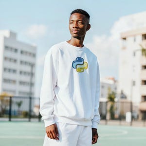 May include: White sweatshirt with a blue and yellow Python logo. The sweatshirt is part of a matching set with white shorts. The person is standing outside on a green surface with buildings in the background.
