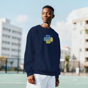 K&ouml;nnte beinhalten: Marineblaues Sweatshirt mit dem Python-Logo in Blau und Gelb auf der Brust. Das Sweatshirt hat einen Rundhalsausschnitt und ist aus einem weichen Material gefertigt. Im Hintergrund sind ein Geb&auml;ude und blauer Himmel zu sehen.