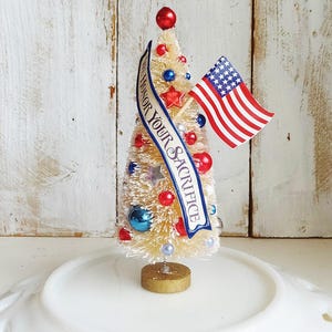 May include: A cream-colored bottle brush tree decorated with red, white, and blue ornaments, stars, and a small American flag. A blue banner draped across the front features the text WE HONOR YOUR SACRIFICE in white capital letters.