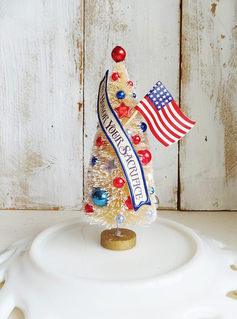 May include: A cream-colored bottle brush tree decorated with red, white, and blue ornaments, stars, and a small American flag. A blue banner draped across the front features the text WE HONOR YOUR SACRIFICE in white capital letters.
