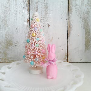 May include: A pink bottle brush tree with pastel-colored beads and a small pink bunny figurine with a pink ribbon. The items are displayed on a white, scalloped-edge plate against a white wooden background.