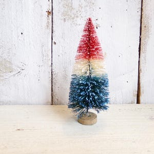 May include: A miniature bottle brush tree featuring a tri-color design with a red top, a white middle section, and a blue base. The artificial tree is constructed with stiff, textured bristles and is mounted on a small, circular wooden base.