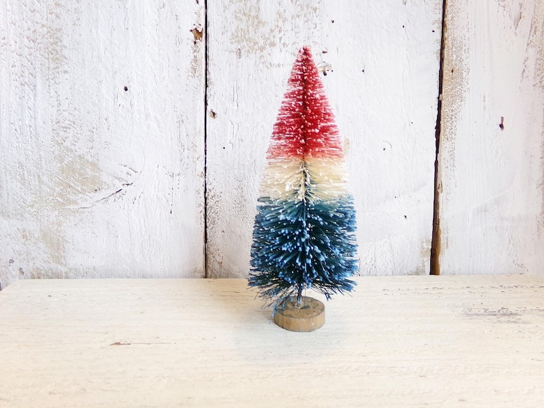 May include: A miniature bottle brush tree featuring a tri-color design with a red top, a white middle section, and a blue base. The artificial tree is constructed with stiff, textured bristles and is mounted on a small, circular wooden base.