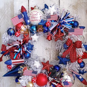 May include: A patriotic wreath featuring a silver tinsel base decorated with red, white, and blue ornaments, including miniature American flags, glittery stars, rocket-shaped accents, and a white bauble with the text God Bless America printed on it.
