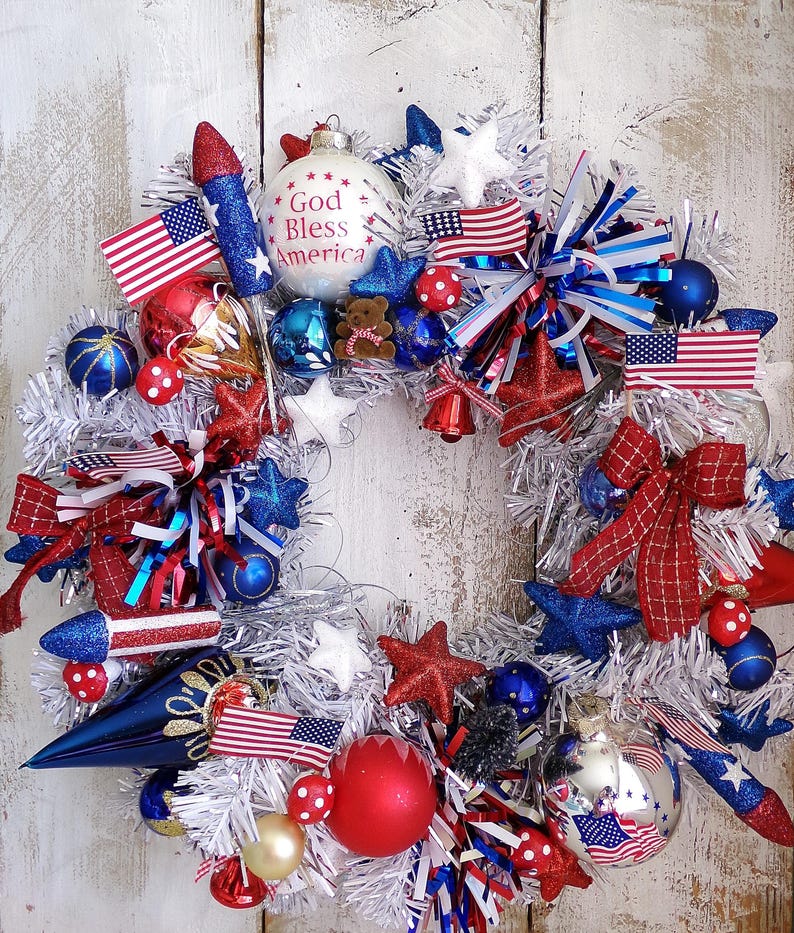 May include: A patriotic wreath featuring a silver tinsel base decorated with red, white, and blue ornaments, including miniature American flags, glittery stars, rocket-shaped accents, and a white bauble with the text God Bless America printed on it.