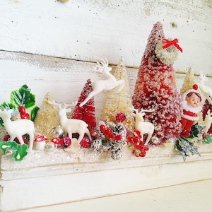 May include: A vintage Christmas display featuring white deer figurines, red bottle brush trees, and a small elf figure. The arrangement includes decorative elements like berries, pinecones, and a small wreath, all set on a white, rectangular base.
