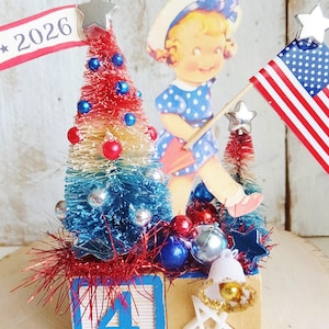 May include: A decorative tabletop display featuring two wooden blocks, one with the number 4, topped with red, white, and blue bottle brush trees, a vintage-style illustration of a child, a 2026 banner, a small American flag, and star accents.