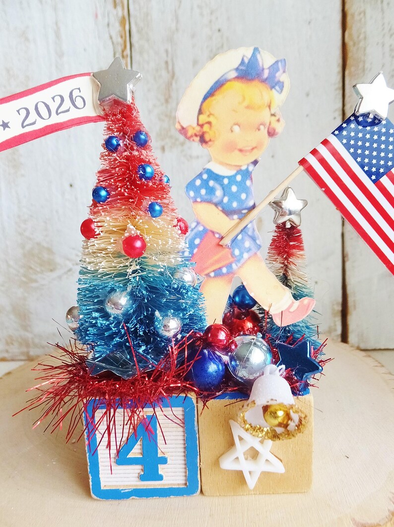 May include: A decorative tabletop display featuring two wooden blocks, one with the number 4, topped with red, white, and blue bottle brush trees, a vintage-style illustration of a child, a 2026 banner, a small American flag, and star accents.