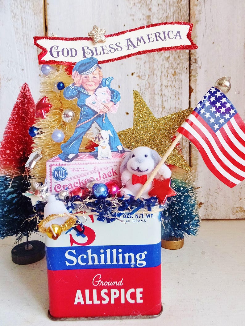 May include: A decorative display featuring a vintage Schilling Ground Allspice tin base, topped with a Cracker Jack box cutout, a sailor boy illustration, a small white plush rabbit, a miniature American flag, and red, white, and blue bottle brush trees.