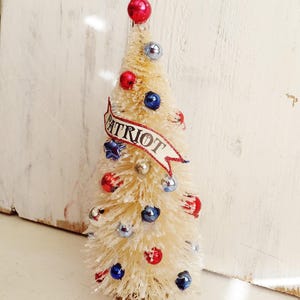 May include: A cream-colored bottle brush tree decorated with small red, white, and blue ornaments. A banner with the word PATRIOT in black text is wrapped around the center. The tree is topped with a single red sphere and has a dusting of white snow.