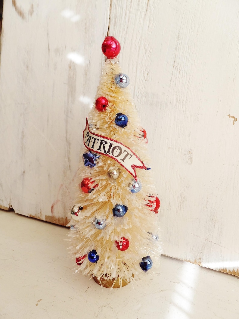 May include: A cream-colored bottle brush tree decorated with small red, white, and blue ornaments. A banner with the word PATRIOT in black text is wrapped around the center. The tree is topped with a single red sphere and has a dusting of white snow.