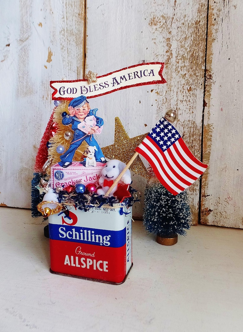 May include: A decorative display featuring a vintage Schilling Ground Allspice tin as a base, topped with a Cracker Jack box, a sailor figure, a small white dog, a gold glitter star, and a miniature American flag with the text God Bless America.