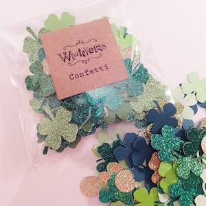 May include: A clear plastic bag filled with assorted green, gold, and navy blue shamrock and circle confetti. A brown paper tag with the words "WhateverS Confetti" is attached to the bag. The confetti is scattered around the bag on a pink surface.
