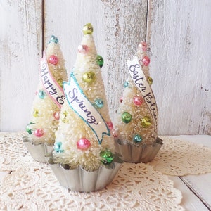 May include: Three white bottle brush trees decorated with colorful beads and ribbons. The ribbons have the text "Happy Spring" and "Easter Blessings".