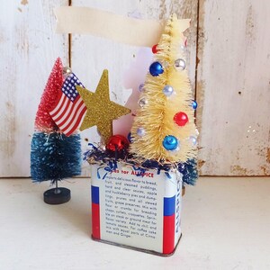 May include: A vintage Allspice tin container decorated with a cream-colored bottle brush tree adorned with red, blue, and silver beads, a gold glitter star, and a small American flag. The tin features red, white, and blue stripes and printed text.