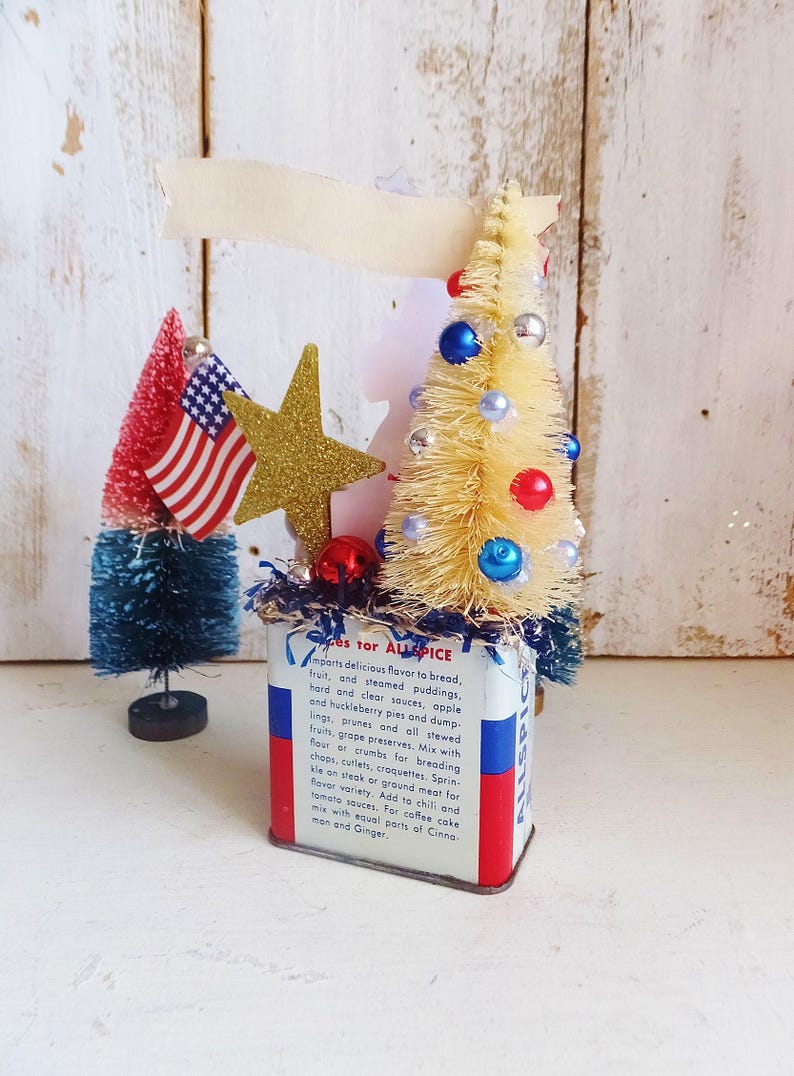 May include: A vintage Allspice tin container decorated with a cream-colored bottle brush tree adorned with red, blue, and silver beads, a gold glitter star, and a small American flag. The tin features red, white, and blue stripes and printed text.