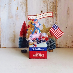 May include: A decorative display featuring a vintage Schilling Ground Allspice tin as a base. It is adorned with a Cracker Jack box, a small American flag, a glittery gold star, a white rabbit figurine, and two miniature bottle brush trees.