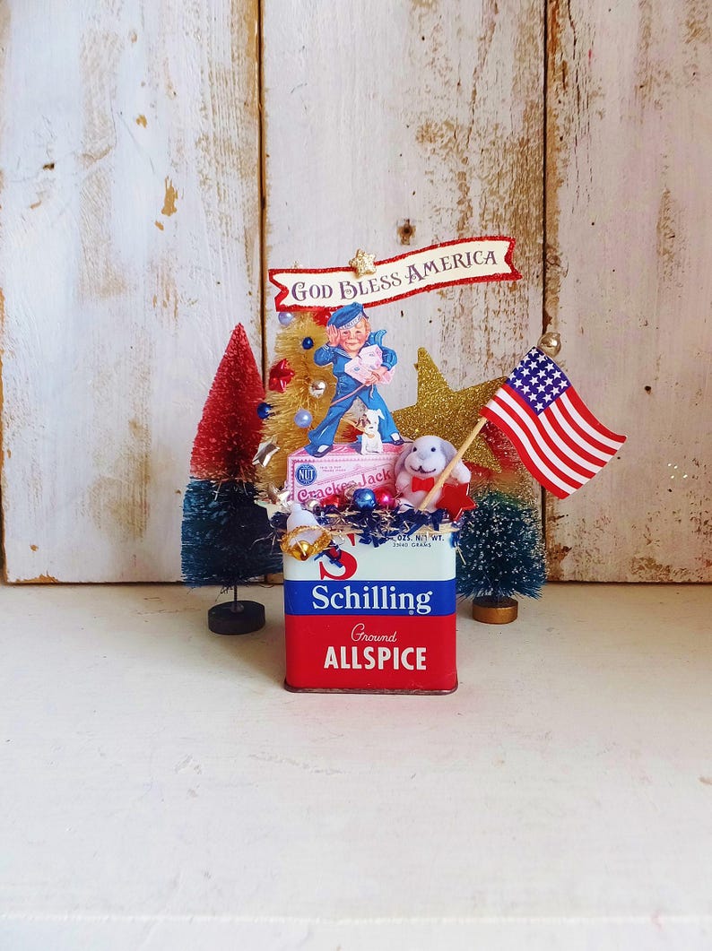 May include: A decorative display featuring a vintage Schilling Ground Allspice tin as a base. It is adorned with a Cracker Jack box, a small American flag, a glittery gold star, a white rabbit figurine, and two miniature bottle brush trees.