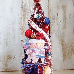May include: A red bottle brush tree decorated with blue and red ornaments, white flowers, and a banner reading AMERICA'S 250TH 1776-2026. A cutout of two children in sailor uniforms stands in front, topped with a silver star and a red base.
