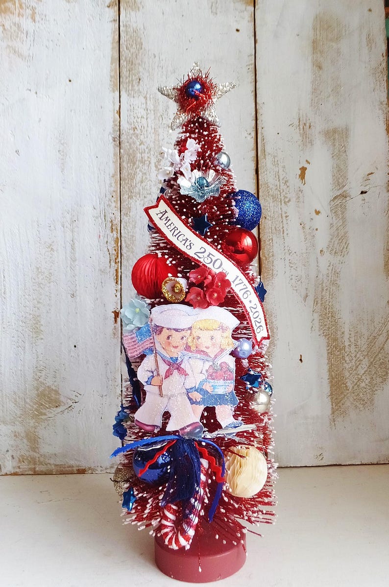 May include: A red bottle brush tree decorated with blue and red ornaments, white flowers, and a banner reading AMERICA'S 250TH 1776-2026. A cutout of two children in sailor uniforms stands in front, topped with a silver star and a red base.
