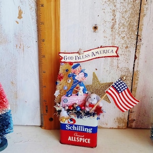 May include: A decorative display featuring a vintage Schilling Ground Allspice tin base, topped with a Cracker Jack sailor figure, a small white dog, a gold glitter star, and a miniature American flag. A banner reads GOD BLESS AMERICA.