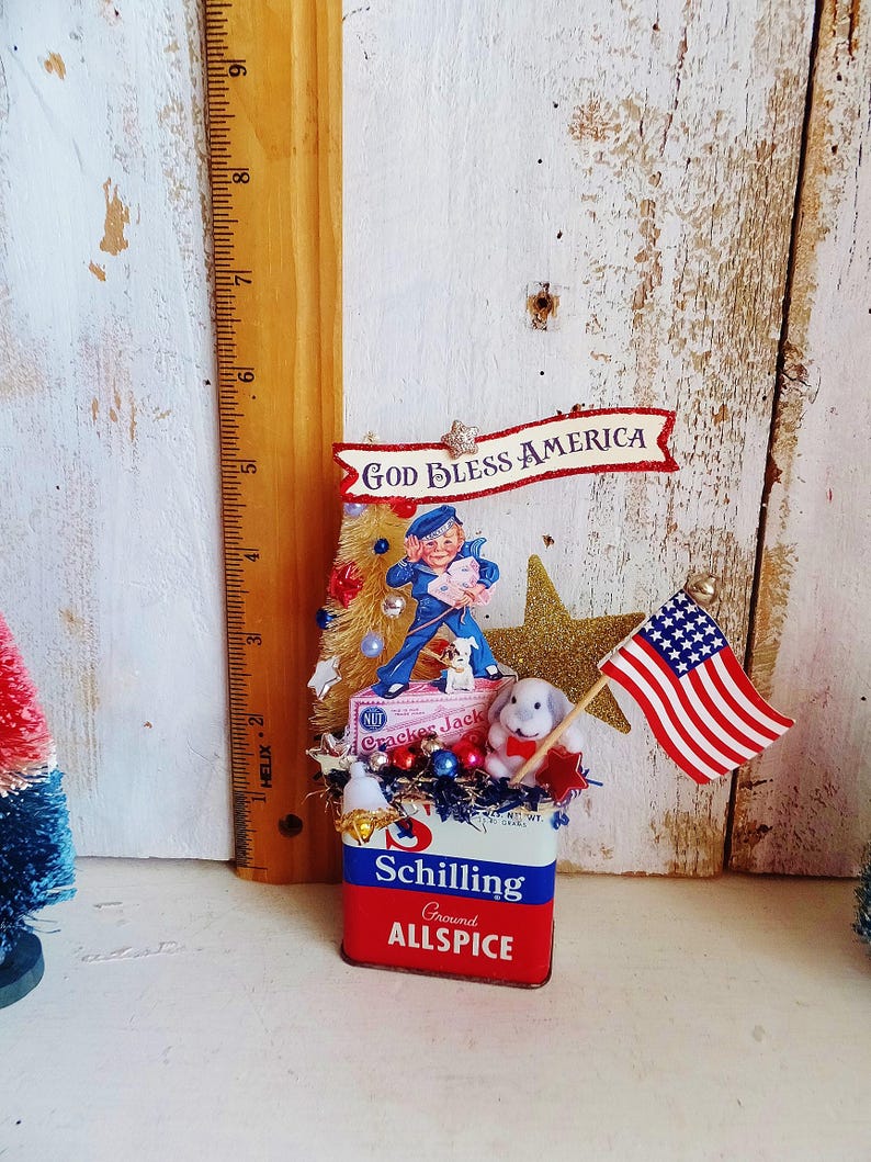May include: A decorative display featuring a vintage Schilling Ground Allspice tin base, topped with a Cracker Jack sailor figure, a small white dog, a gold glitter star, and a miniature American flag. A banner reads GOD BLESS AMERICA.